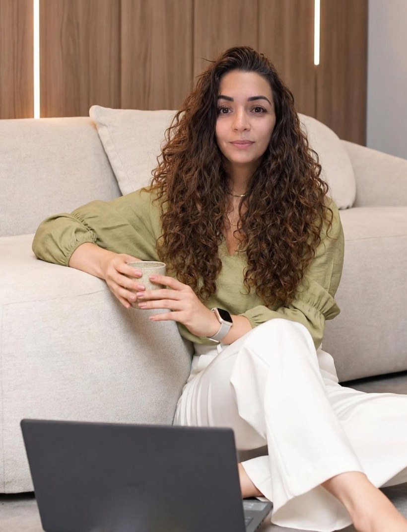 Jeniffer Jiménez sentada en el suelo, recargada en un sillón claro, con una taza entre las manos y una laptop y libros frente a ella.